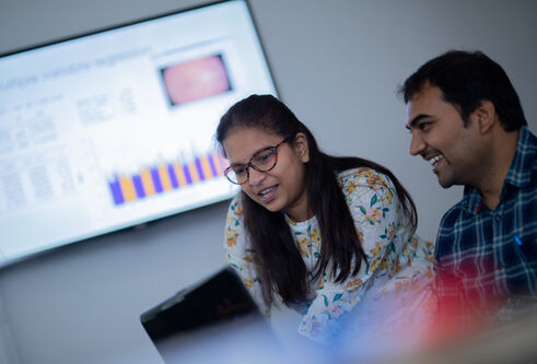 Students compare data on a laptop with a large screen behind them showing charts
