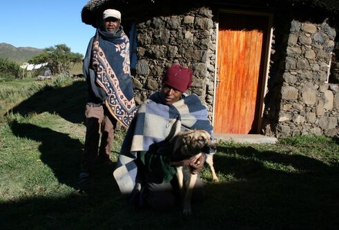 2 people and a dog sitting outside of a stone house with a wooden door