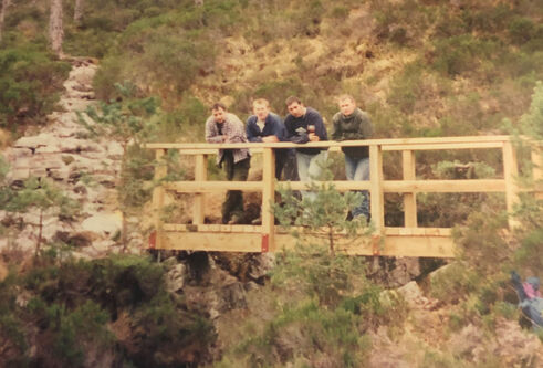 Four men in their twenties lean against the handrail on a freshly built, unpainted, wooden bridge. The bridge crosses a small rocky gorge surrounded by foliage and further rock formations.