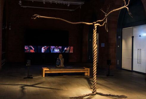 rope twisted to look like a tree, in a dark gallery with a screen showing people playing stringed instruments