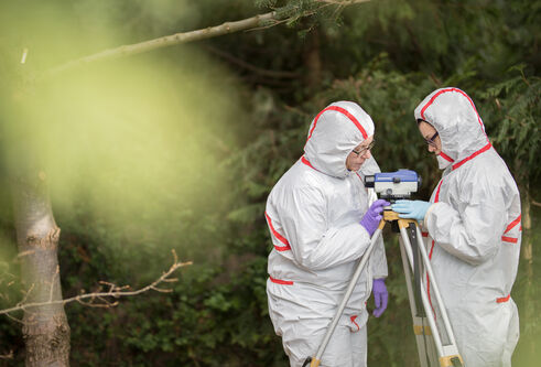 two people in full body clean suits look at a cameralike device on a tripod in the woods