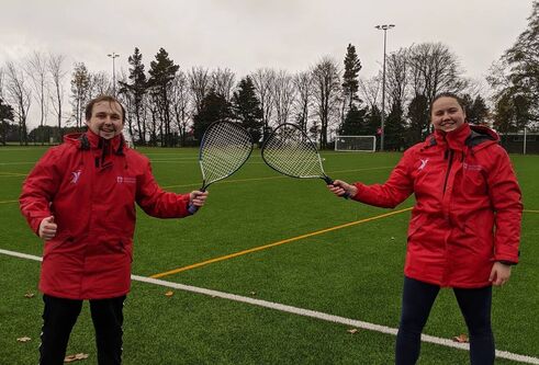 full shot of president dan and club member wearing red rain jackets standing on green grass pitch holding tennis rackets