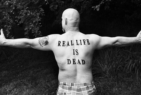 naked man with outstretched arms and tattoo of 'real life is dead'