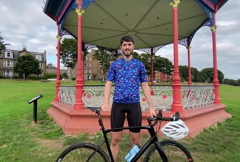 male in early 20's wearing tight blue and black cycling wear posing with black frame bike, standing in front of bandstand with red posts, surrounded by vibrant green grass