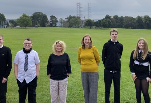 a group of five schoolchildren wearing uniform standing each side of two middle aged women in a park of vibrant green grass