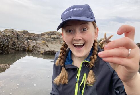 Close up of young woman in 20's sporting braided blonde hair, wearing a navy cap and jumper, excitedly holding up a small starfish against a background of water