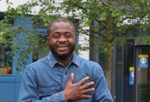 young black male in 20's wearing light blue shirt, posing with smile in front of green hedges and trees