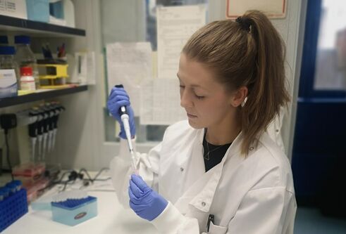 woman in lab coat and blue gloves holding pipette, background is a laboratory