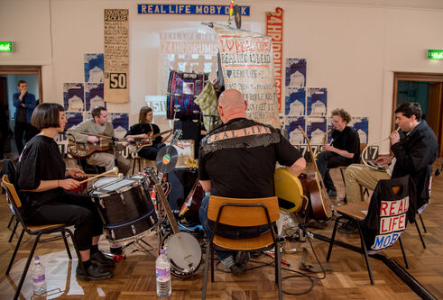 orchestra in a small room surrounded by posters with text 'Real Life Moby Dick'