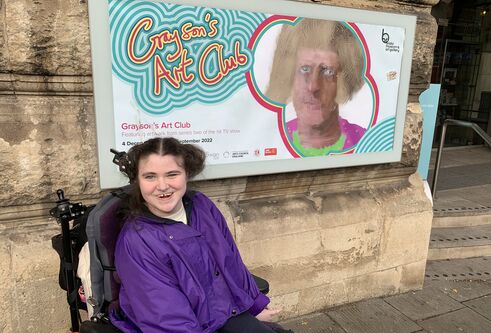 A young girl wearing a bright purple jacket, sat in a motored wheelchair, excitedly smiling in front of exhibition poster