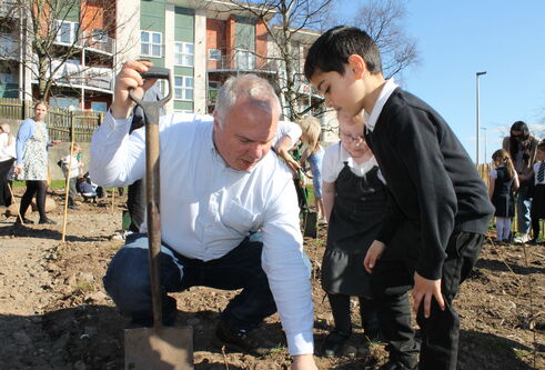 middle aged man crouched down on muddy land, balancing using a shovel, pointing to the ground while a young boy keenly looks on 