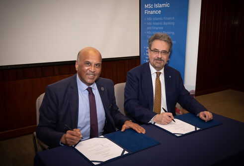 two middle aged men, dressed formally in a suit and tie, sat beside one another at table both signing two pieces of white documents