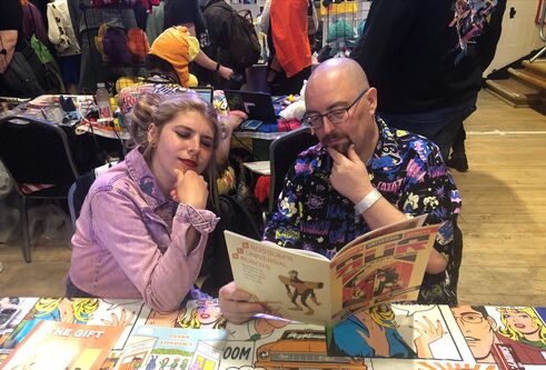 Two people sit at a table covered with comics, both looking together at an open comic with their hands on their chins.