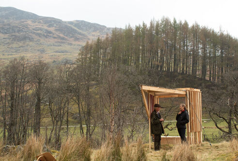 two people standing under a wooden structure in the middle of a grassy area with bare trees and rolling hills in background