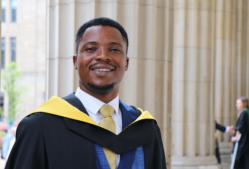 young black man stands in graduation robes, posing against large stone pillars