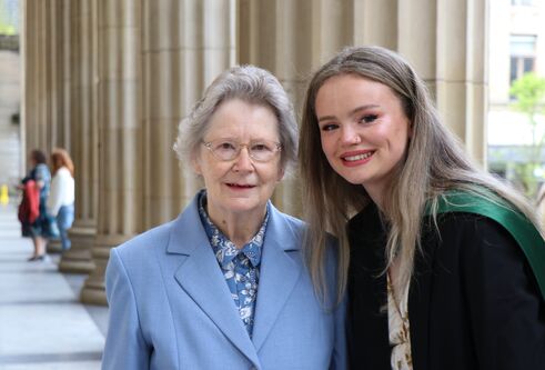 elderly woman wearing blue blazer standing next to young blonde woman wearing graduation grown
