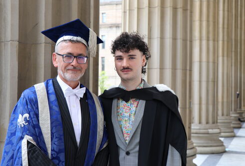 a middle aged man wearing a blue gown and tassled hat stands next to a young man in his 20's wearing a black graduation robe