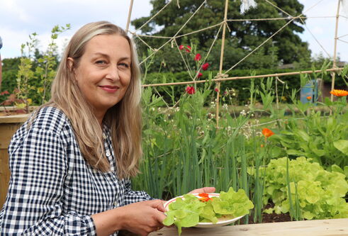 a woman with blonde hair, wearing a grey checkered shirt, holding a plate full of green salad beside a planter full of sprouting green plants 