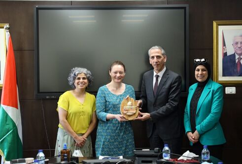 Jane Macaskill stands holding an award, surrounded by three Palestinian colleagues. They are standing in an office space with the flag of Palestine hanging beside a desk.