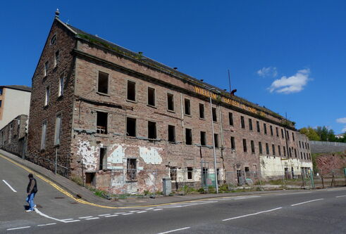 front view of a large derelict jute mill building on the edge of an empty road. The building is in a state of disrepair, with dark window openings on three different levels