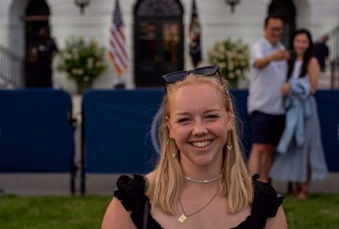 A young blonde girl in her early 20's, wearing a black short sleeved shirt, stands on grass in front of the White House. The American and Presidential Seal flags are in the background
