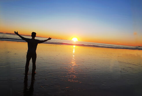 Person standing on beach at sunset with hands raised