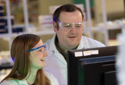 Two forensic scientists in lab equipment smiling as they view a computer screen.