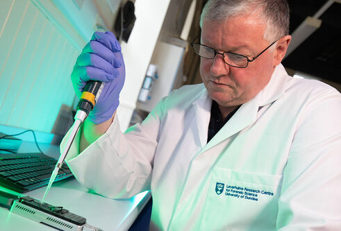 A male scientist in a lab coat and gloves pipetting a solution into a MinION, a pocket-sized device used sequence DNA & rNA.