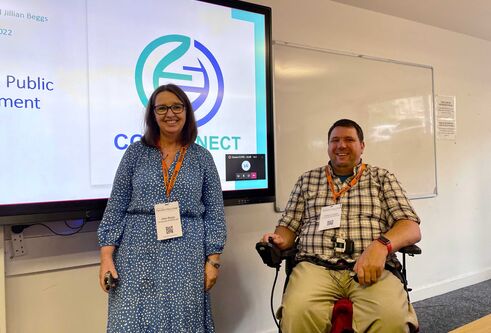 A woman in a blue dress stands next to a man in a wheel chair, in front of a presentation displayed on a screen.