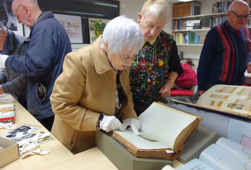 People are standing around a set of tables, a woman in the middle is leaning over looking at a book.