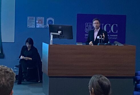 A man standing at a presentation stand, and a woman sits taking notes beside him.