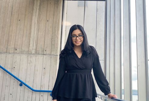 A woman standing in a stairwell by some windows, wearing a black dress.