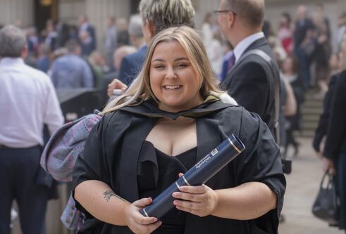 Smiling graduate with long hair, wearing gown and holding certificate tube