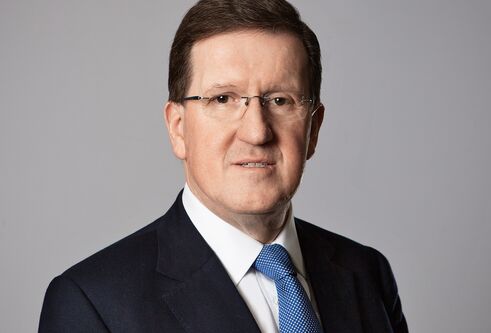 Man with brown hair and glasses wearing a dark suit with a white shirt with a blue tie is looking at the camera