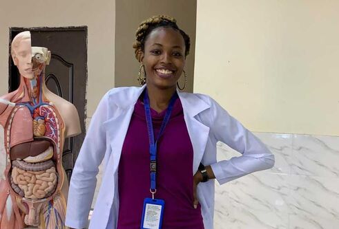 A woman wearing a purple dress and blazer, stands with her left hand on her hip. Anatomical models are behind her.