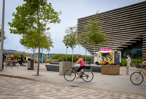People sat a picnic tables outside V&A Dundee as cyclists go past