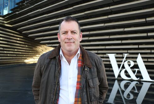 A man in a white shirt and brown jacket standing outside infront of the V&A museum