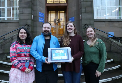 Four people stoof outside infront of steps, holding a plastic box with a blue and green label on it
