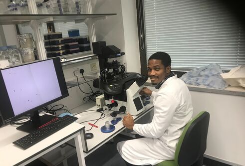 A man wearing a white lab coat sits at a desk with a computer and other gadgets.