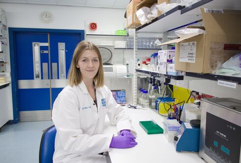 Woman wearing white lab coat, sat at a table wearing purple gloves, in front of shelves containing science equipment