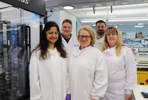 Group of scientists wearing lab coats and protective eyewear, three woman in front and two men in row behind, standing in a lab with science equipment around them.