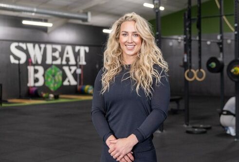 woman with long, wavy blond hair looking at camera wearing a black long sleeved top, with gym equipment in the background