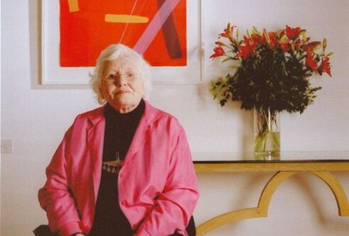Wilhelmina Barns-Graham sits in front of a red print and bunch of flowers, wearing a pink blazer.