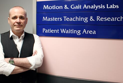 A man wearing a white shirt and black waistcoat stands next to a blue sign.