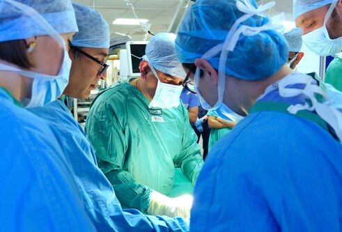 Surgeons wearing blue and green clothing, masks and hair nets, stand around looking at a demonstration together.