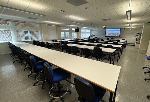 A room full of desks and blue chairs with wheels. There are windows along the left wall.
