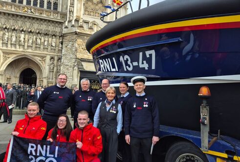 Lifeboat outside Westminster Abbey, with a group of people standing to one side of it. Front row of three people wearing red outfits hold a flag while six people behind wear dark uniforms and medals.