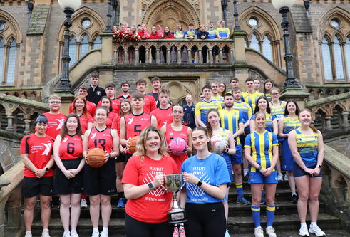 Large group of people wearing sports kit standing on outside steps, one half wearing red kits and the other half wearing blue and yellow striped kits. Front two women hold a trophy between the two of them.