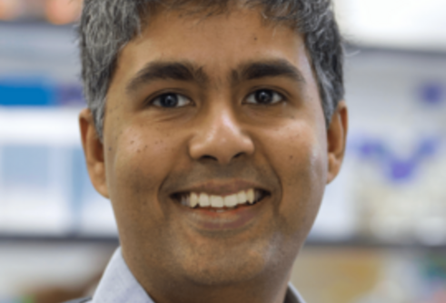 man in a blue shirt and grey jumper is stood in a lab smiling at the camera
