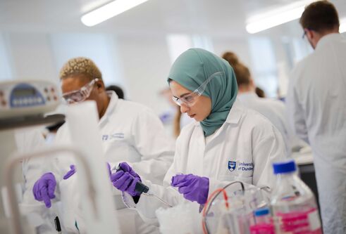 Two females in lab coats, purple gloves and safety glasses. One girl is holding an instrument in her hand and looking at it.